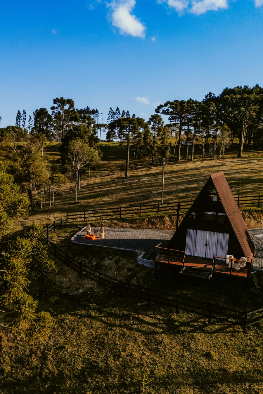 Vista aérea do chalé A-frame cercado por araucárias sob céu azul