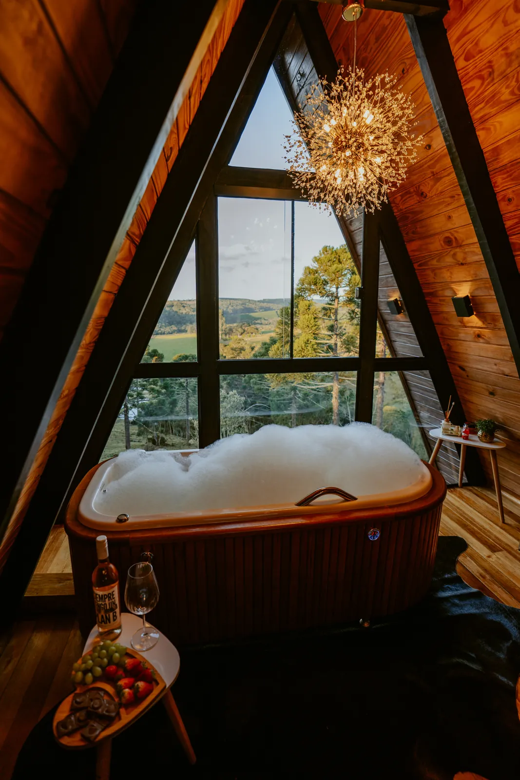 Banheira de hidromassagem sob o vitral triangular do chalé, com lustre e vista para o vale